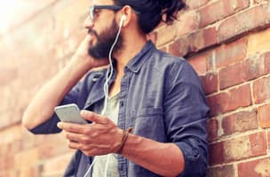 A man is using a mobile phone with wired headphones in his ears, while leaning against a brick wall