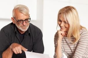 A man with white hair and a beard points at a letter being held by a woman in a beige and white striped top