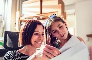 woman with short red bobbed hair in a blue and white striped long-sleeved top reading mail next to a young girl with long brown hair with sunglasses on top of her head