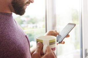 man in a burgundy top with black-rimmed glasses, standing to one side, holding a mug on his phone