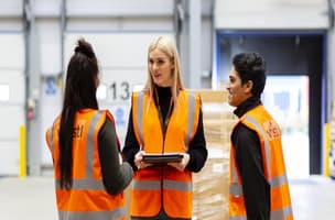 Three Whistl warehouse operatives in orange hi-vis jackets are discussing fulfilment elements, they operatives are stood in front of loading dock doors