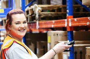 A Whistl Fulfilment employee smiles to the camera next to warehouse shelving full of eCommerce products. 