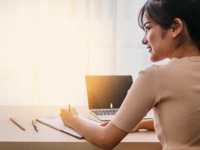 A woman is facing an open laptop in front of a window, with the sun shining through.