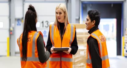 Three Whistl warehouse operatives in orange hi-vis jackets are discussing fulfilment elements, they operatives are stood in front of loading dock doors