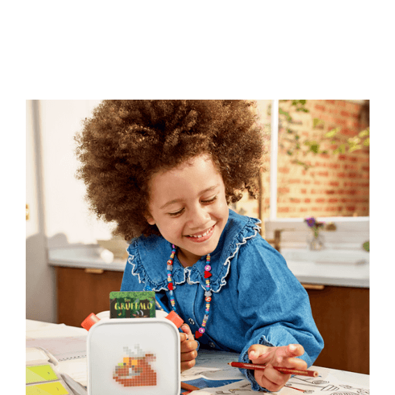 A young child smiles while drawing with a Yoto player in front of her with the Gruffalo card showing